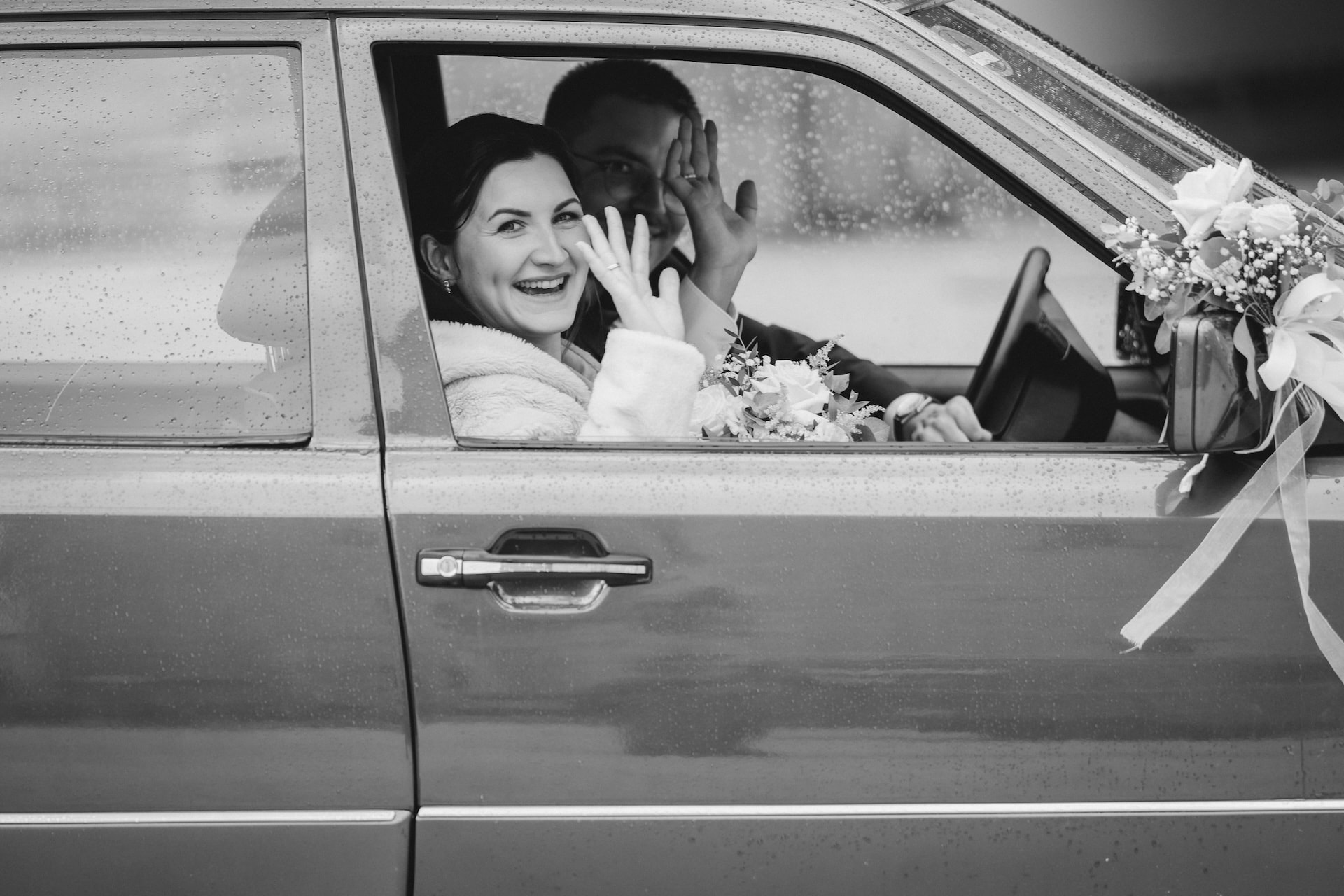 Hochzeitsfotografie Wiblingen, Braut zeigt lachend Ehering aus dem Autofenster im Regen, Schwarzweißfoto